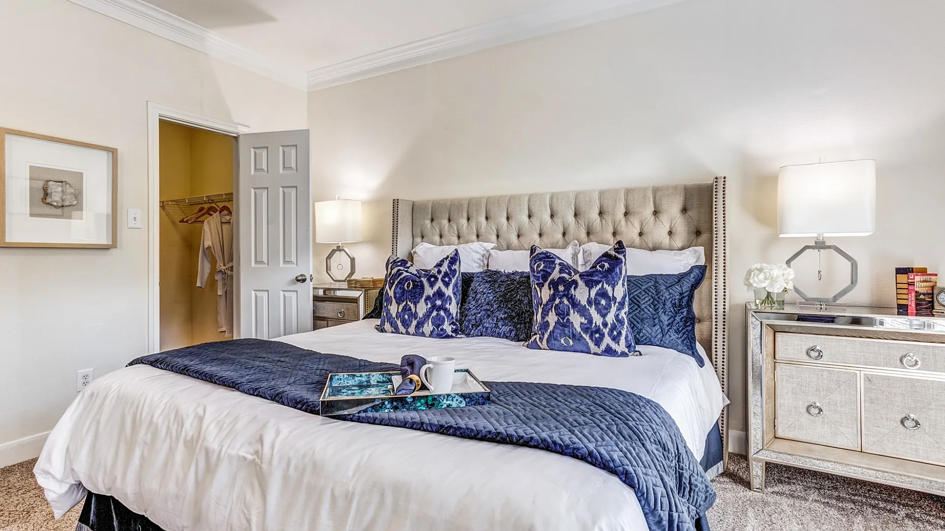 Elegant bedroom with a plush king-sized bed, stylish blue accents, and modern decor featuring a mirrored nightstand and ambient lighting.