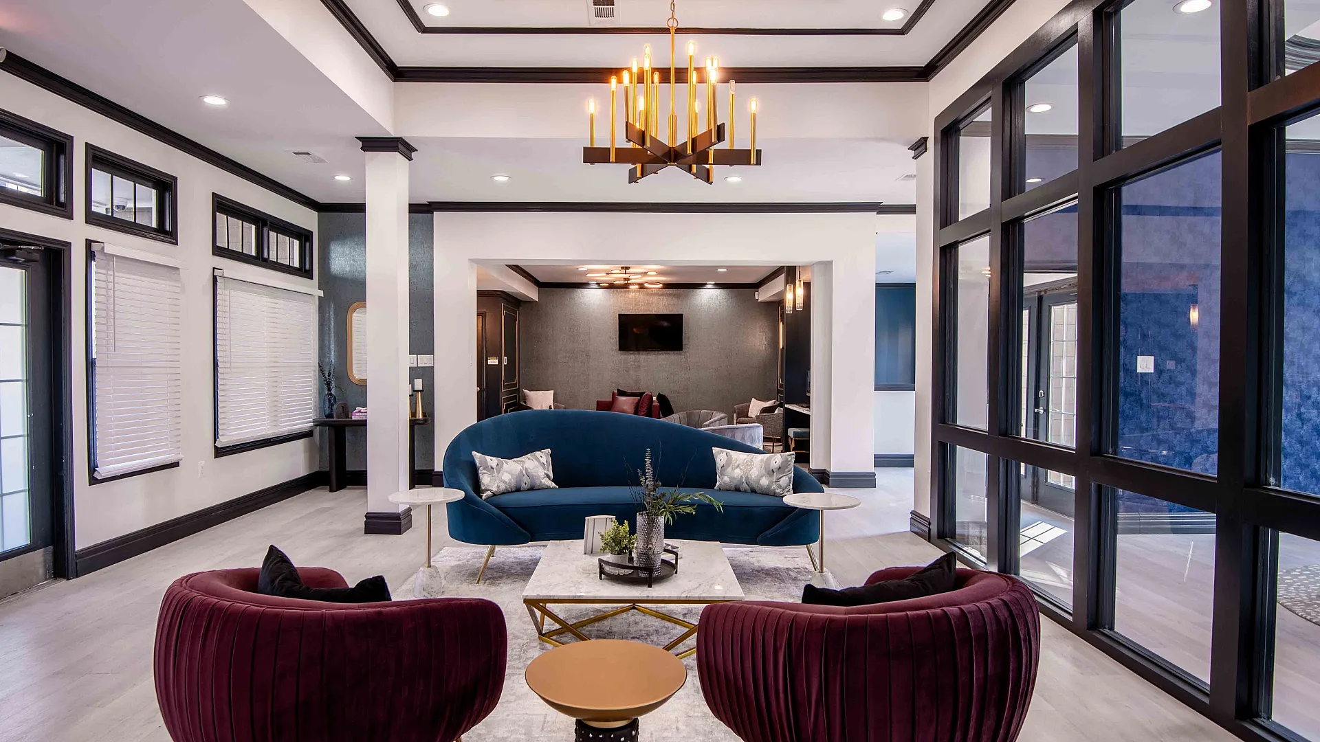 Elegant living room with plush burgundy chairs, a stylish blue sofa, modern chandelier, and large window panels offering natural light.