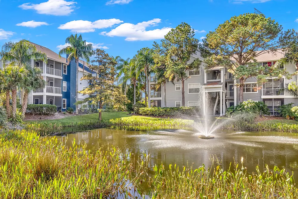 Charming apartment complex surrounded by lush greenery and a serene pond with a central fountain, perfect for tranquil living.