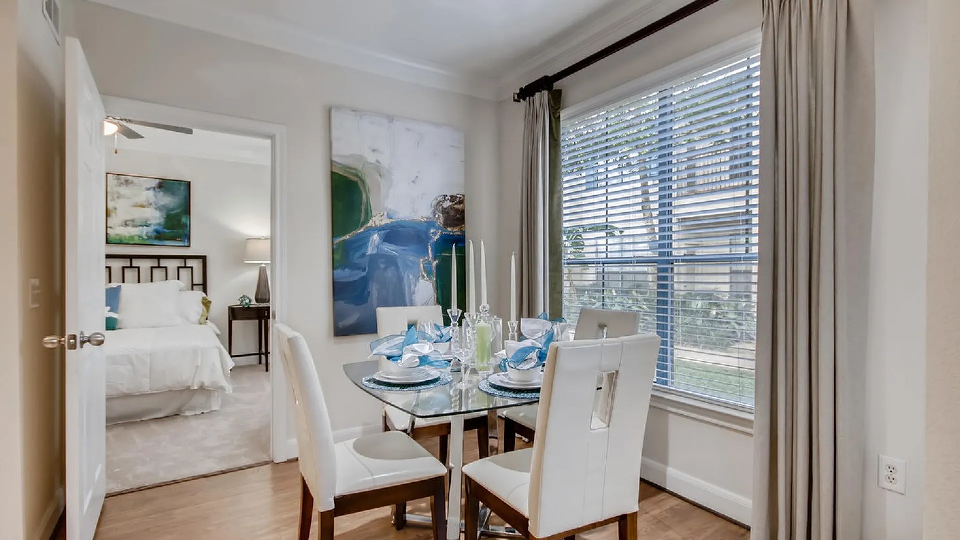 Bright dining area with elegant table setting and large window, leading into a cozy bedroom with modern decor.