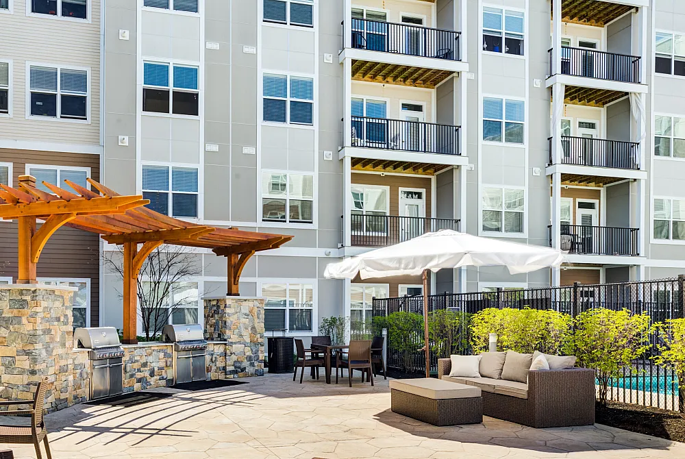 Modern apartment complex exterior with stylish balconies, outdoor grilling area under a pergola, and inviting patio seating with lush greenery.