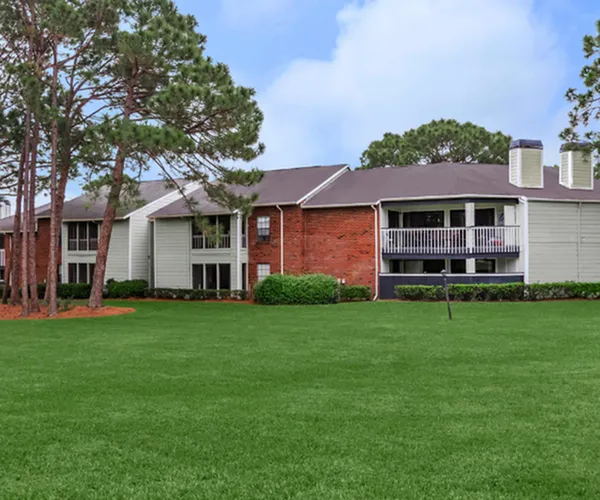Charming apartment complex with red brick facade, two-story units, and lush green lawn surrounded by tall trees, perfect for serene living.