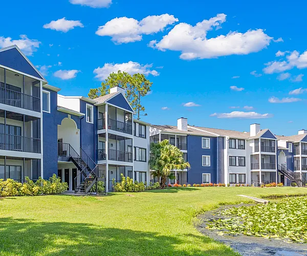 Charming apartment complex with modern blue and white design, surrounded by lush landscaping and a tranquil pond under a bright blue sky.
