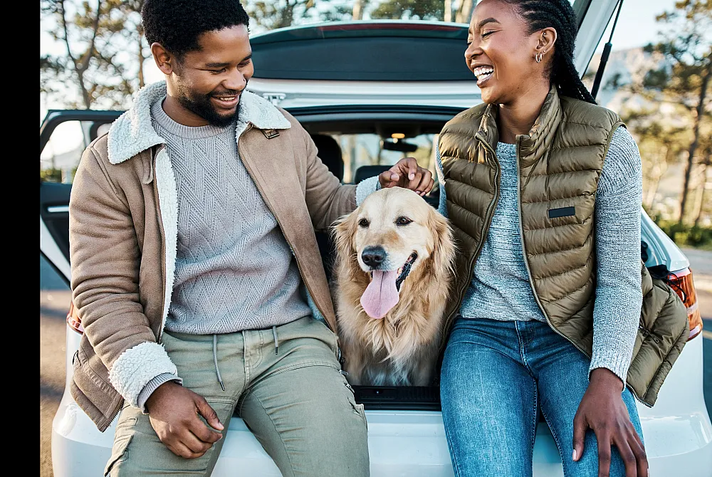 Joyful couple with a golden retriever sitting at the back of an open car, enjoying a sunny day outdoors.