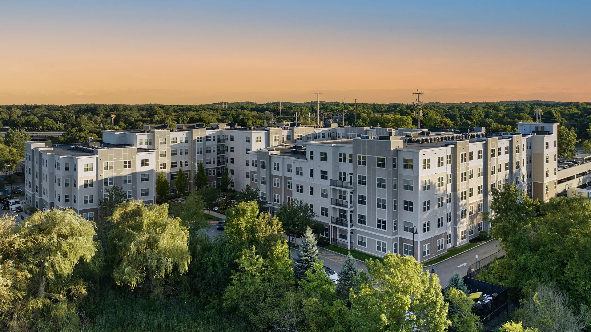Modern apartment complex in a lush, green setting at sunset, featuring multiple stories and sleek architectural design.
