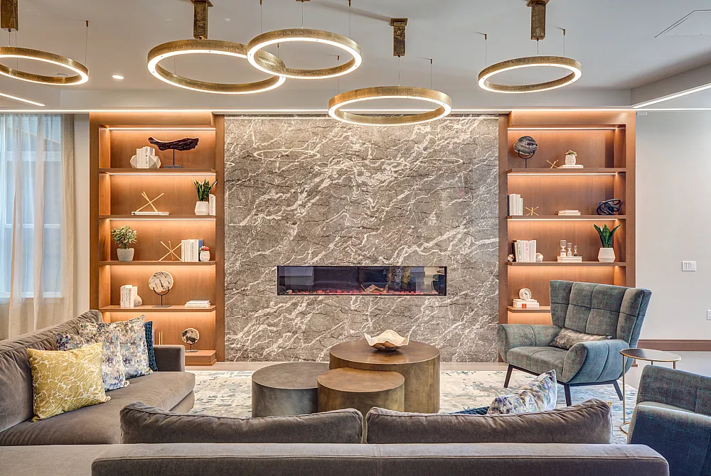 Elegant lounge with modern furnishings, featuring a chic gray marble fireplace, stylish shelving, and luxurious seating illuminated by golden ring lights.