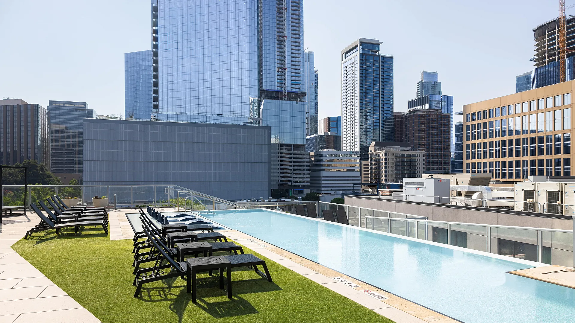 Sleek rooftop infinity pool with modern city skyline views, surrounded by stylish lounge chairs and lush green turf.