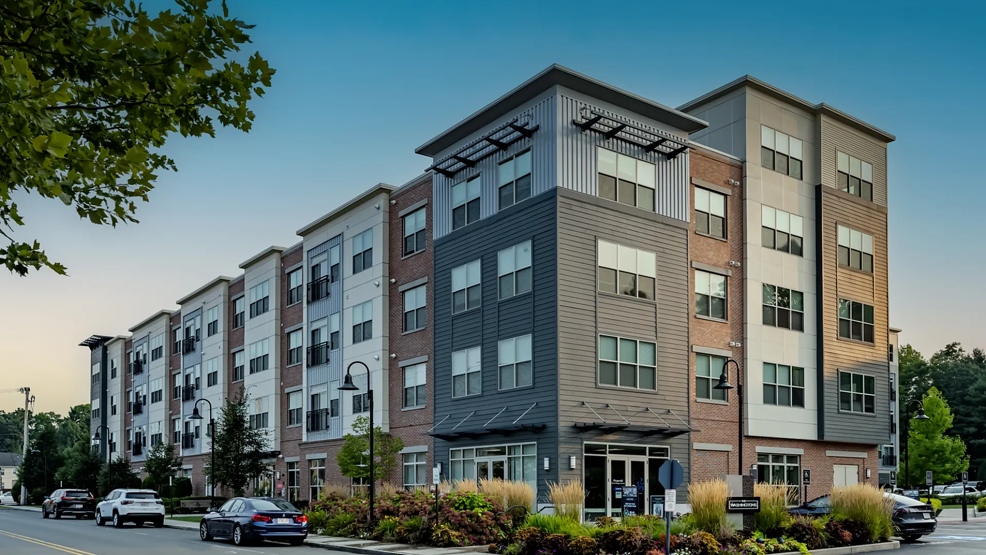 Modern multi-story apartment building with sleek design, surrounded by lush landscaping and street parking, under a clear blue sky.