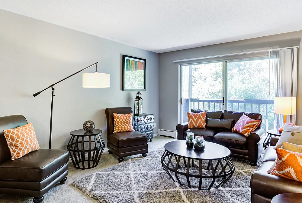 Inviting living room with stylish leather seating, vibrant orange cushions, and large window opening to lush greenery.