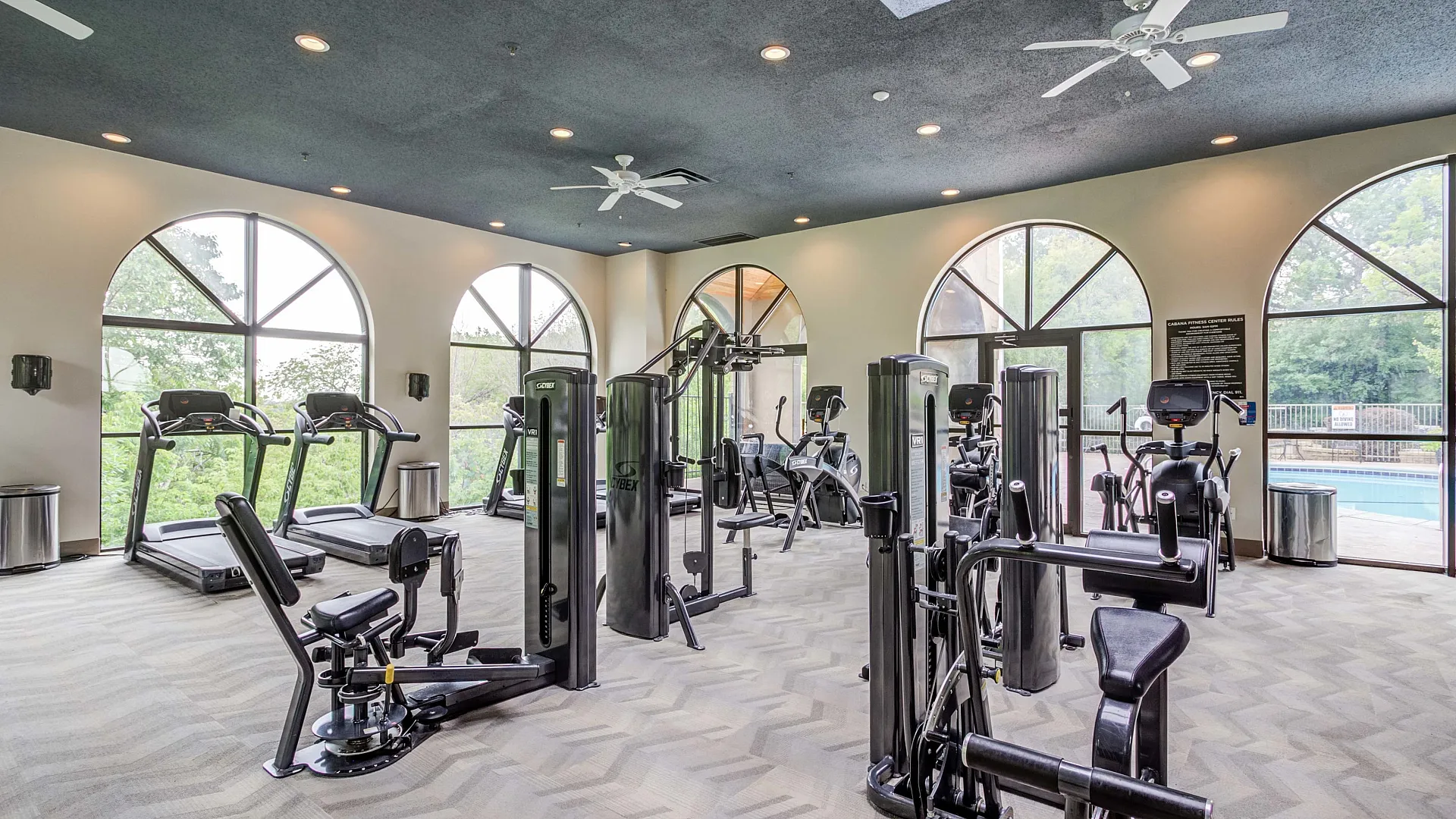 Bright and airy fitness center with modern exercise equipment and large arched windows overlooking lush greenery and a pool.