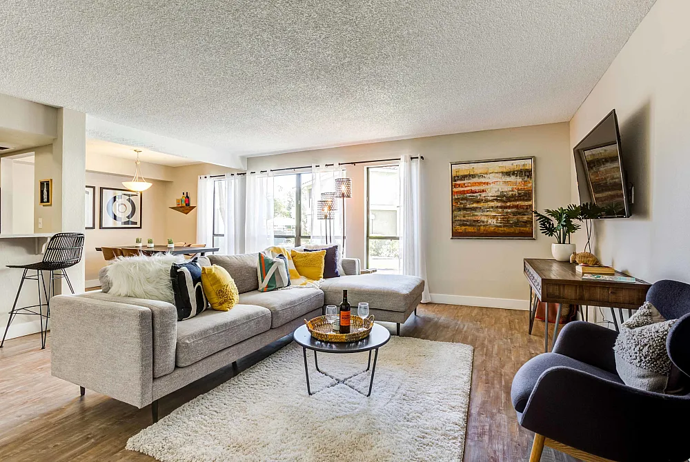 Bright and inviting living room with modern decor, featuring a sleek sectional sofa, vibrant throw pillows, and a wall-mounted TV, complemented by large windows and elegant artwork.