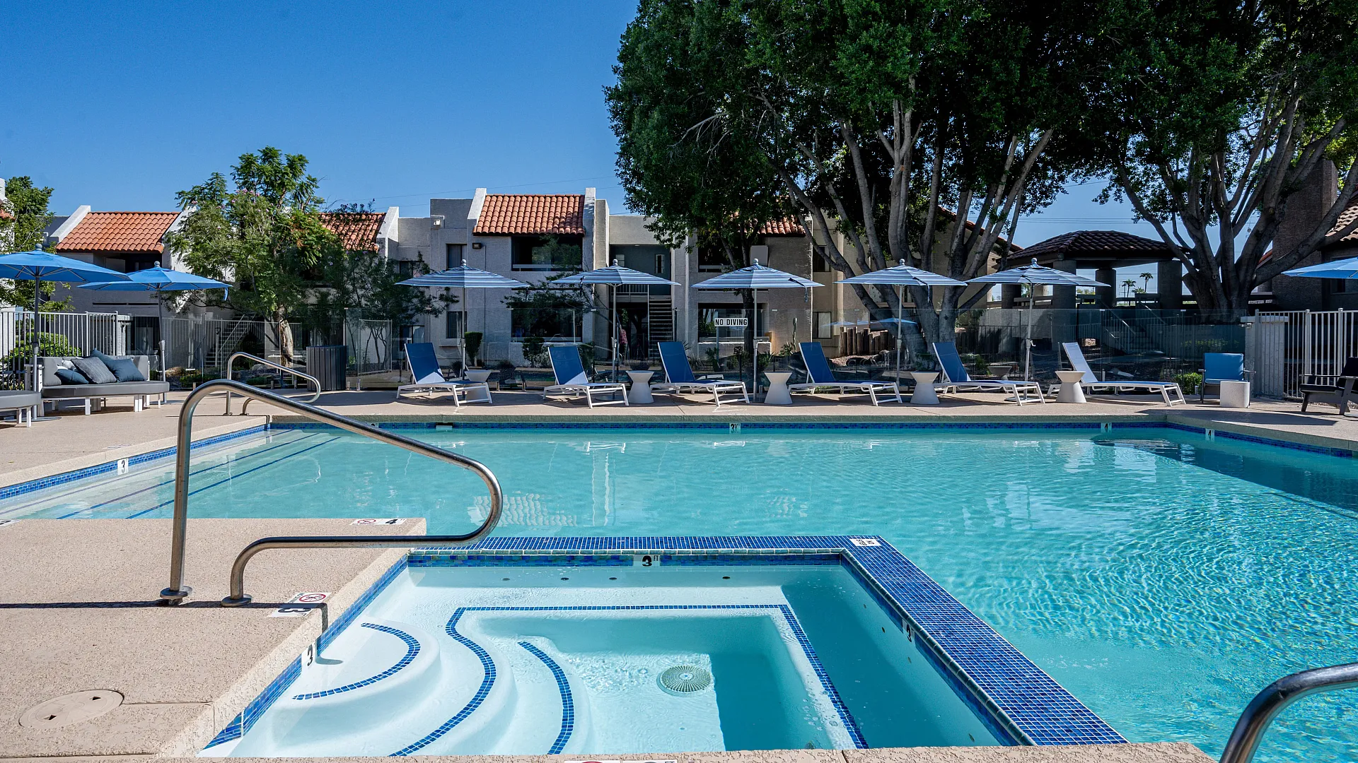 Inviting swimming pool with sun loungers and umbrellas, surrounded by lush trees and charming residential buildings.
