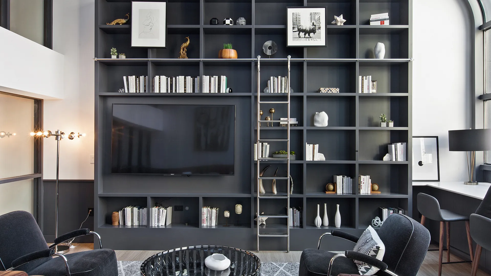 Chic living room with sleek shelving, integrated TV, stylish armchairs, and a modern ladder for a contemporary aesthetic.