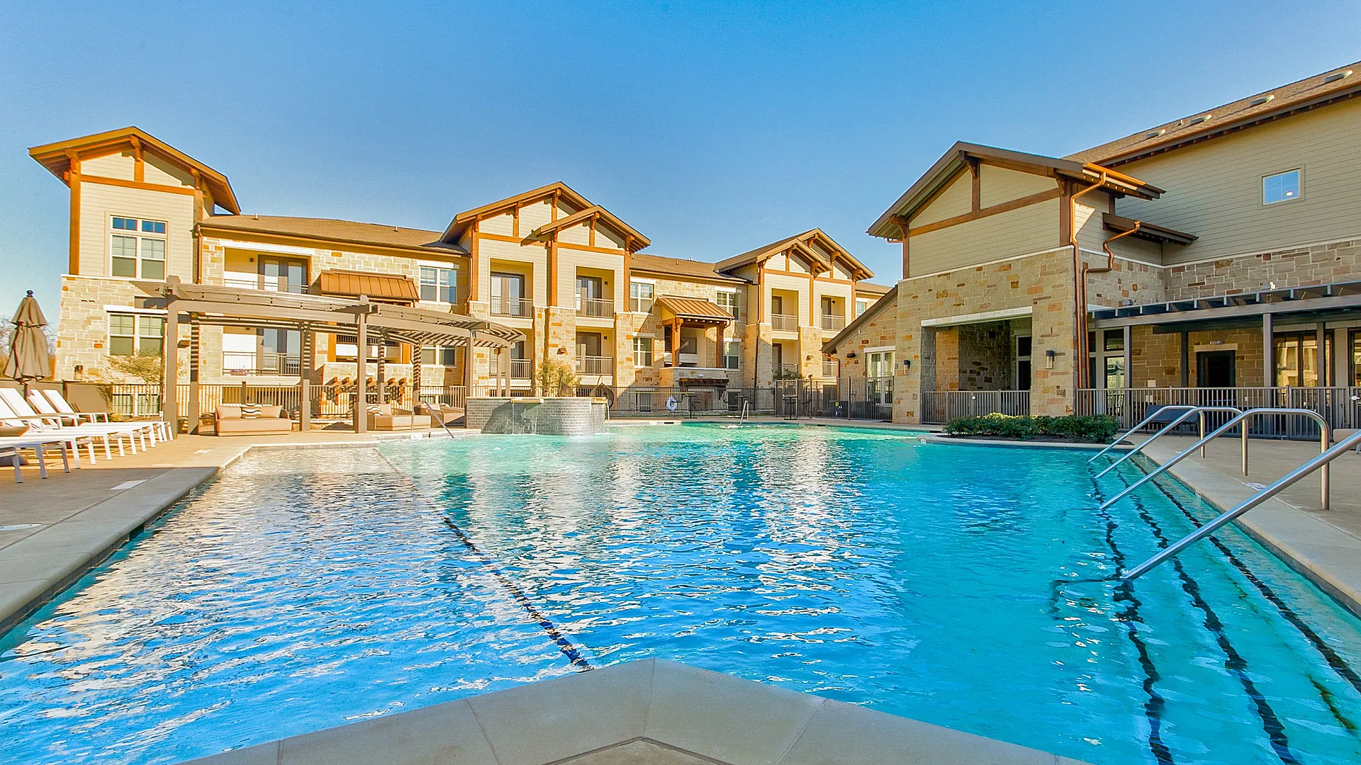 Luxurious apartment complex with a stunning outdoor pool, sun loungers, and elegant stone architecture under a clear blue sky.