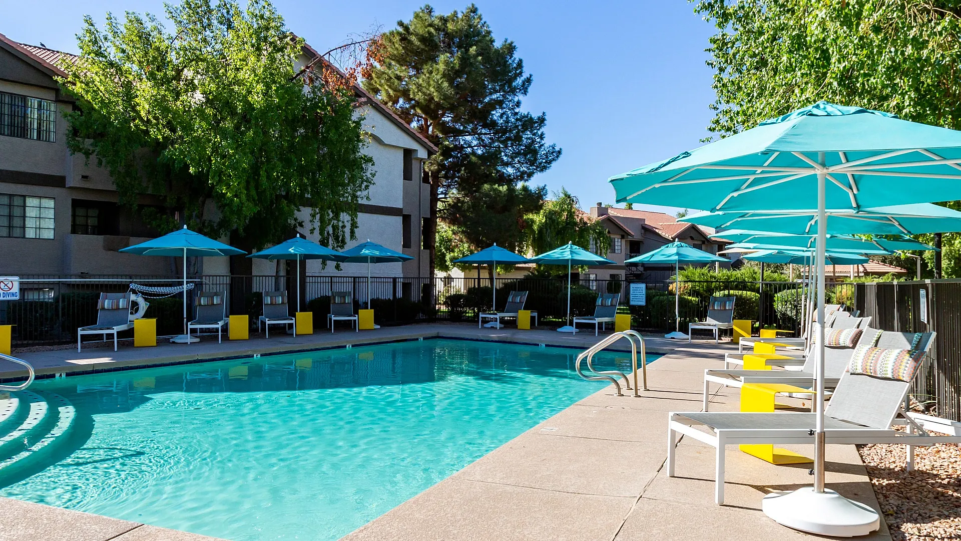 Inviting apartment complex pool area with turquoise umbrellas, sun loungers, and lush surrounding greenery under a clear blue sky.