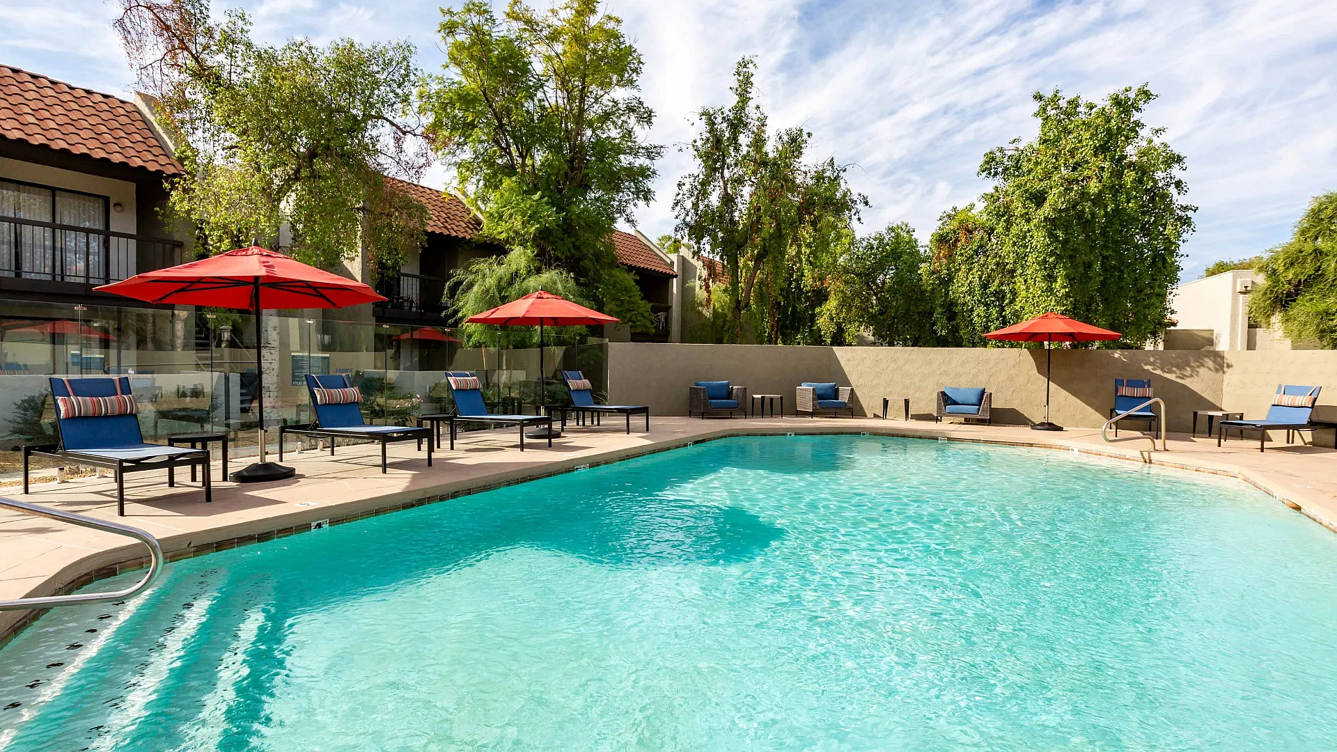 Inviting outdoor pool area with red umbrellas and comfortable lounge chairs, surrounded by lush greenery and charming multi-level buildings.
