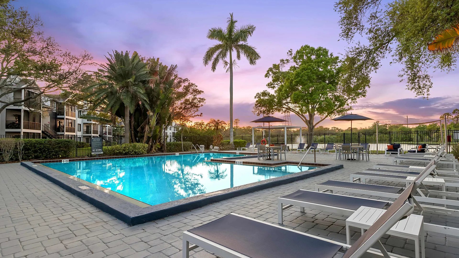 Inviting pool area with lounge chairs and umbrellas set against a stunning sunset backdrop, surrounded by lush greenery and modern apartment buildings.