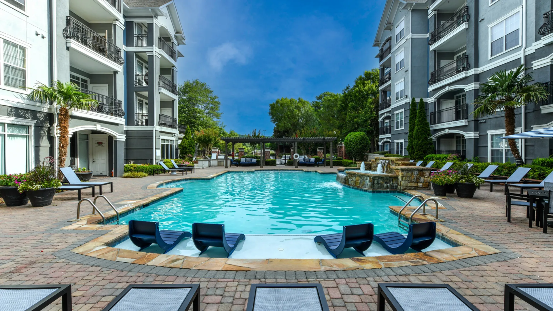 Luxurious apartment pool with elegant loungers, surrounded by lush greenery and stylish multi-story buildings.