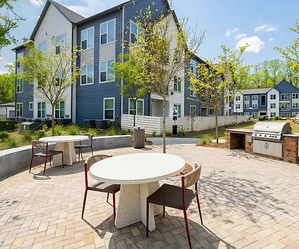 Charming outdoor communal area with modern round tables and a built-in grill, set against a backdrop of contemporary apartment buildings and lush landscaping.