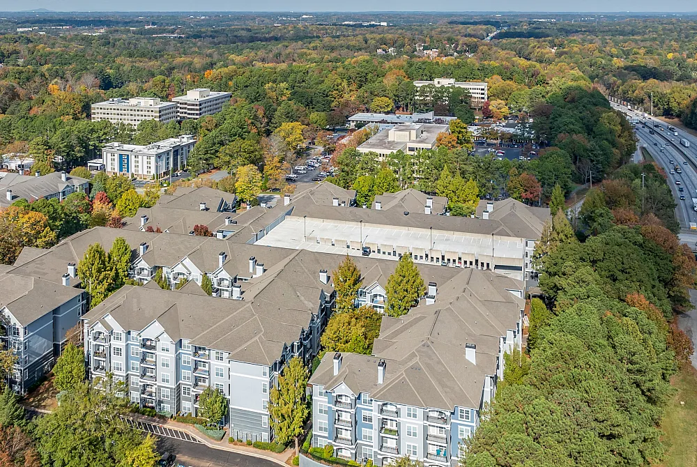 Aerial view of a modern apartment complex surrounded by lush greenery and vibrant autumn foliage, offering scenic living with convenient highway access.