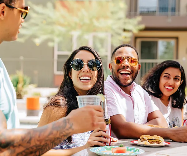Group of people enjoying a sunny day, laughing and sharing drinks at an outdoor patio.