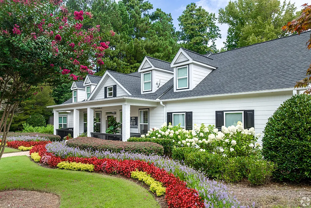 Charming suburban home with a manicured garden featuring vibrant flowers and lush greenery, surrounded by tall trees and bright blue sky.