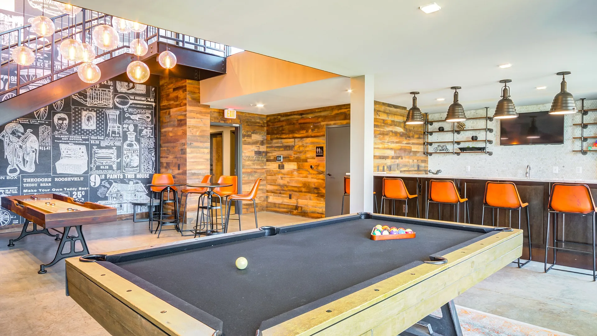 Bright and modern recreation area featuring a stylish pool table, trendy orange bar stools, and a chic chalkboard wall under elegant pendant lighting.