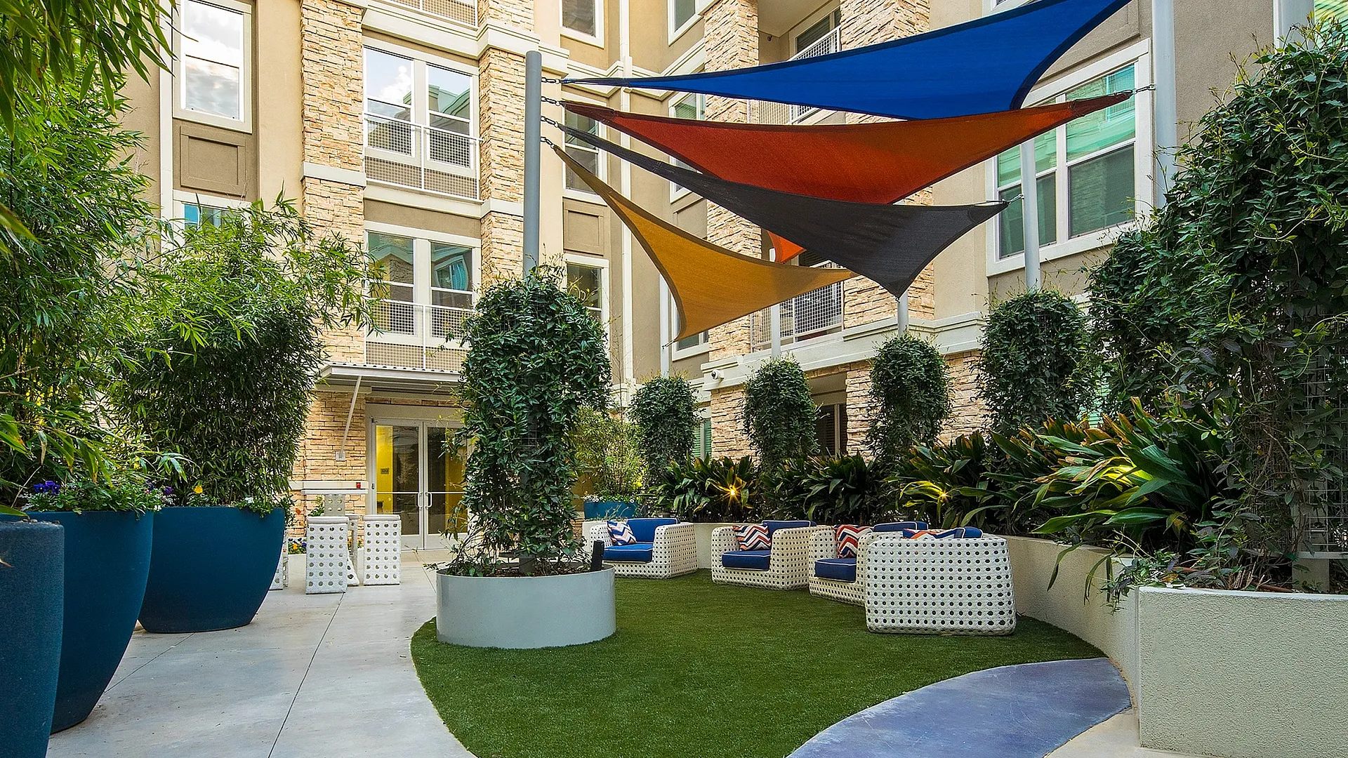 Vibrant apartment courtyard featuring colorful shade sails, modern outdoor seating, and lush green landscaping.