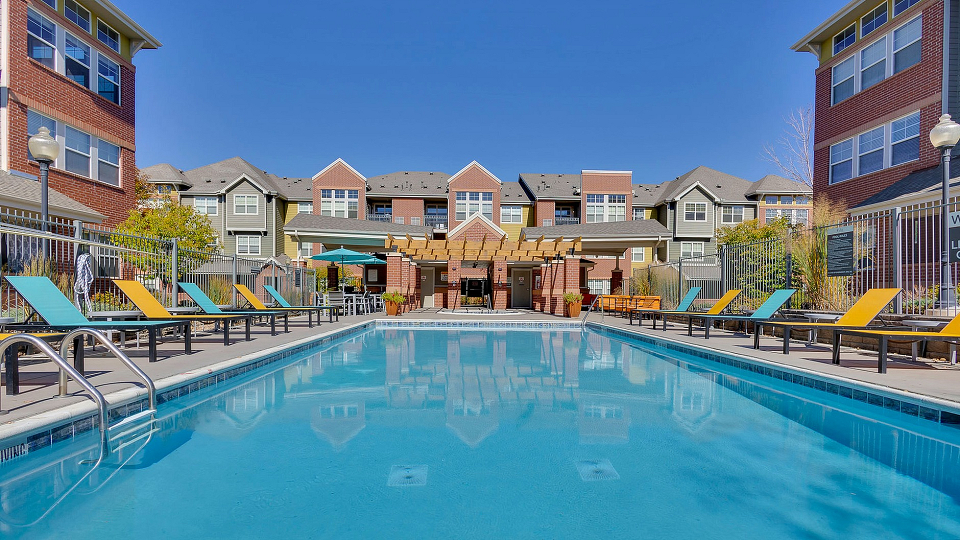 Charming apartment complex with vibrant poolside ambiance, featuring colorful lounge chairs and elegant multi-story buildings under a clear blue sky.