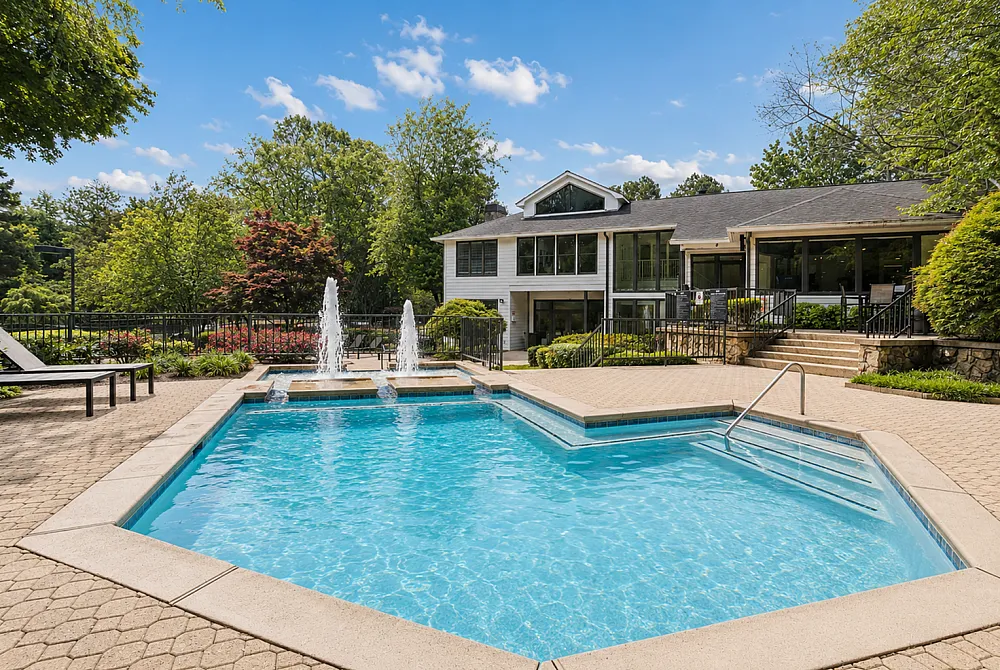 Luxurious apartment complex with a sparkling swimming pool, elegant fountains, and lush green surroundings under a clear blue sky.