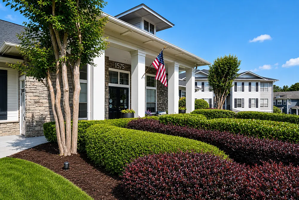 Charming apartment complex entrance with lush landscaping, stone facade, and vibrant greenery under a clear blue sky.