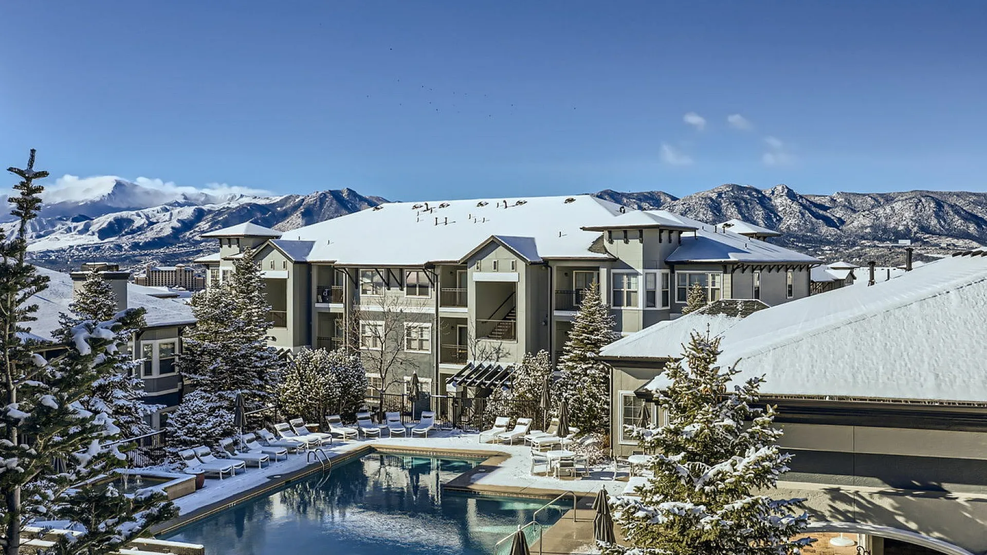 Breathtaking winter view of a luxurious apartment complex with a snow-covered pool area, surrounded by stunning mountain scenery.