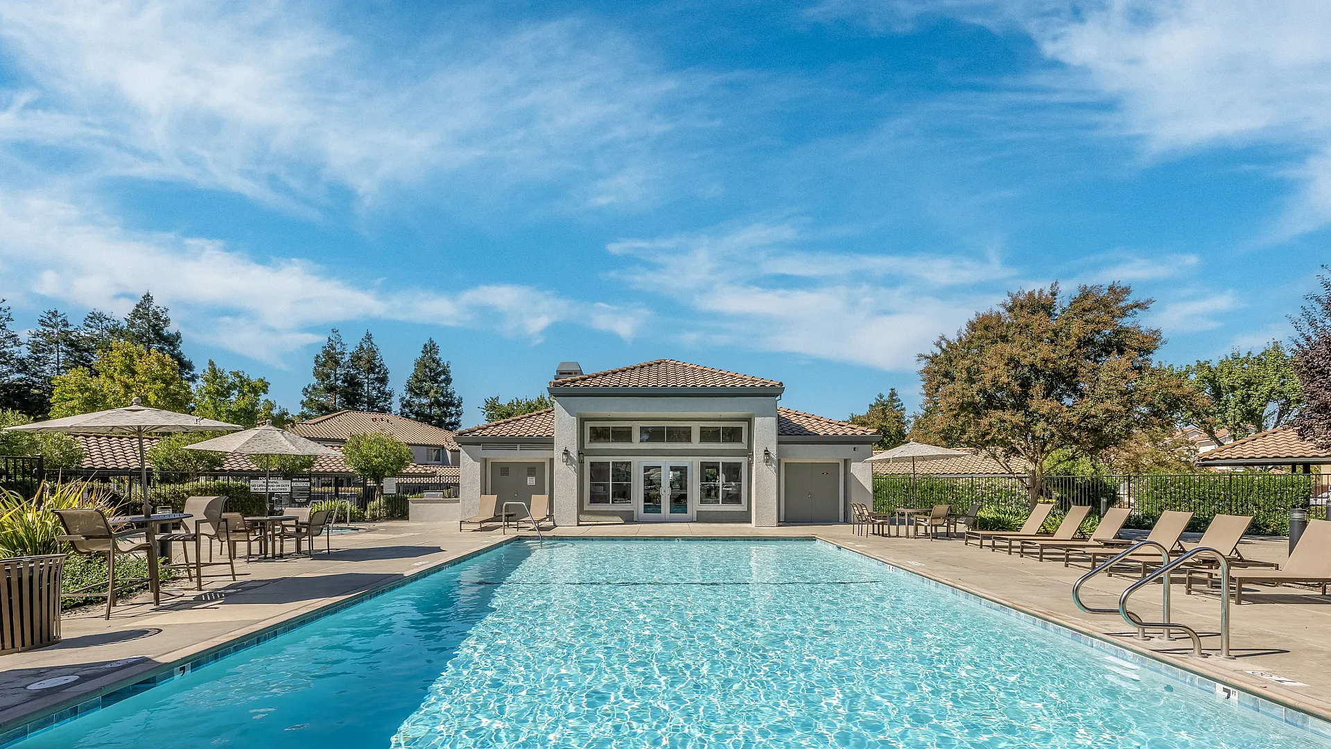 Luxurious community pool area with elegant lounge chairs and umbrellas, surrounded by lush greenery and modern architecture under a bright blue sky.