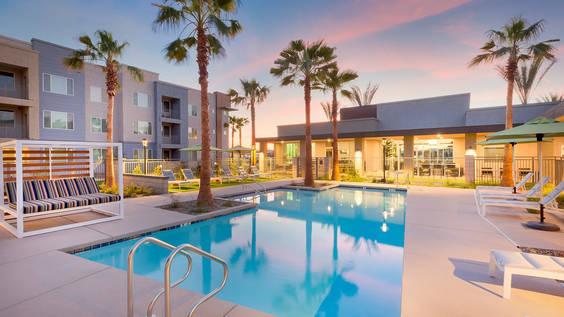 Elegant apartment complex with a stunning palm-lined pool, modern poolside lounge, and serene sunset view.