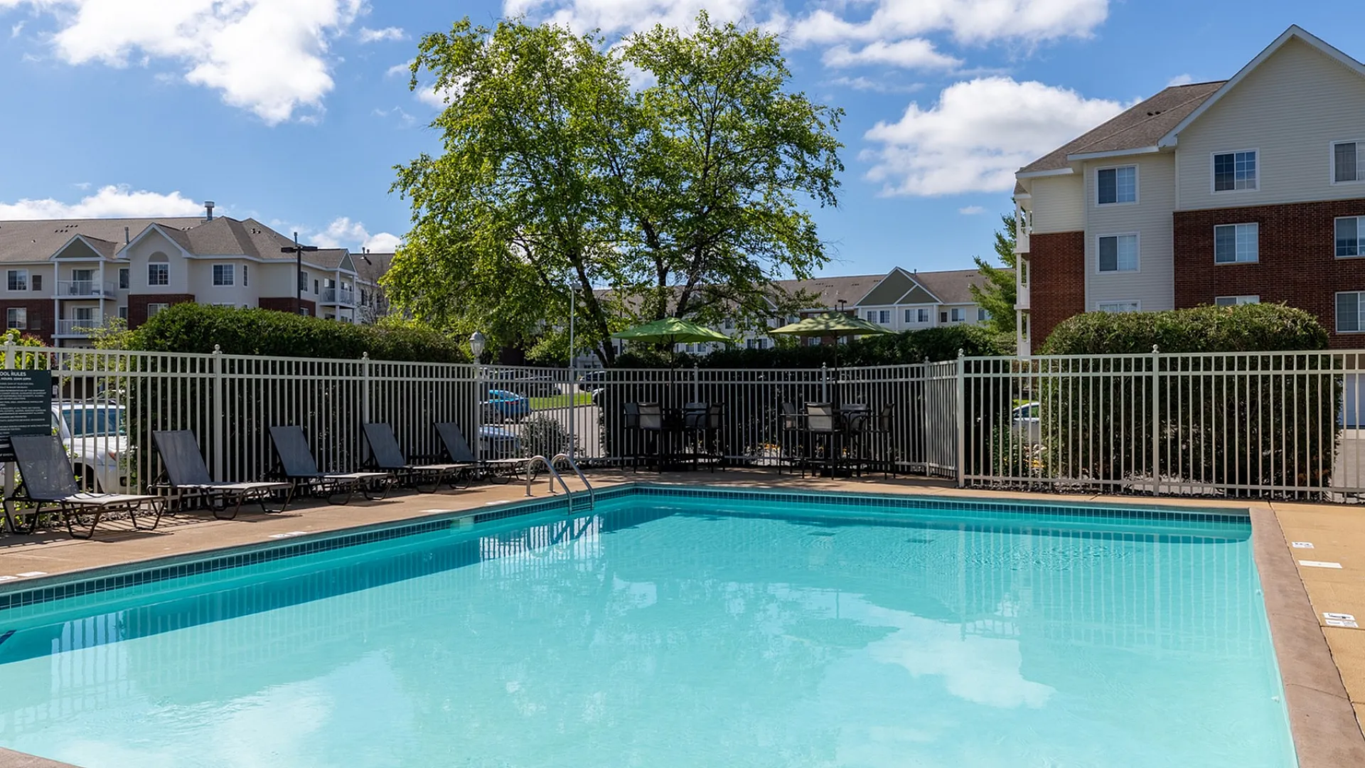 Inviting outdoor pool area with comfortable lounge chairs, surrounded by lush greenery and modern apartment buildings under a clear blue sky.