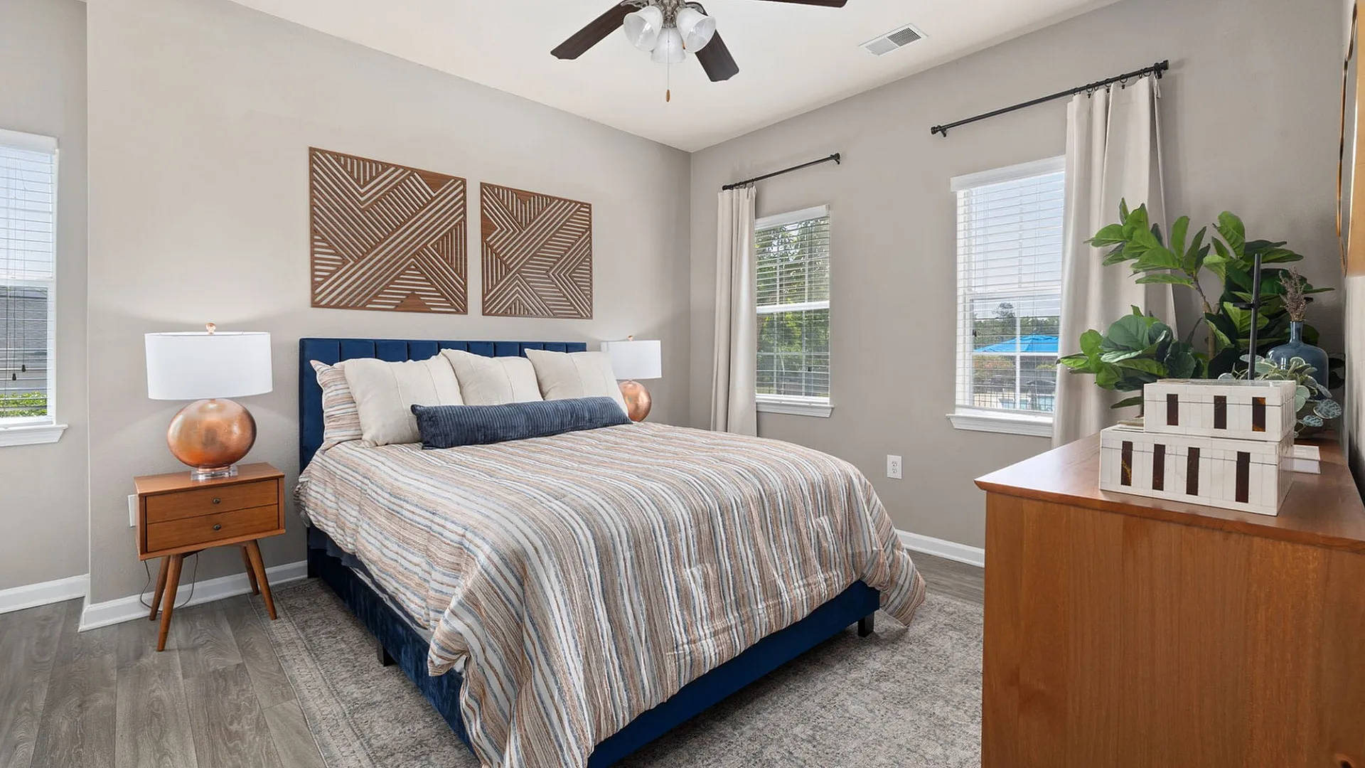 Inviting bedroom with modern decor, featuring a stylish bed with a striped comforter, wooden nightstand, and large windows offering natural light.