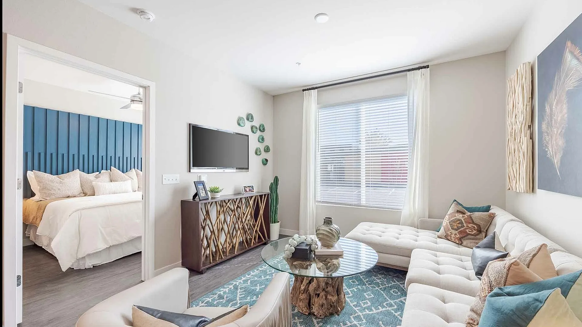 Bright and modern living room with elegant decor, featuring a comfortable L-shaped sofa, vibrant pillows, and a sleek glass coffee table. Adjacent bedroom with stylish blue accent wall creates a harmonious flow.