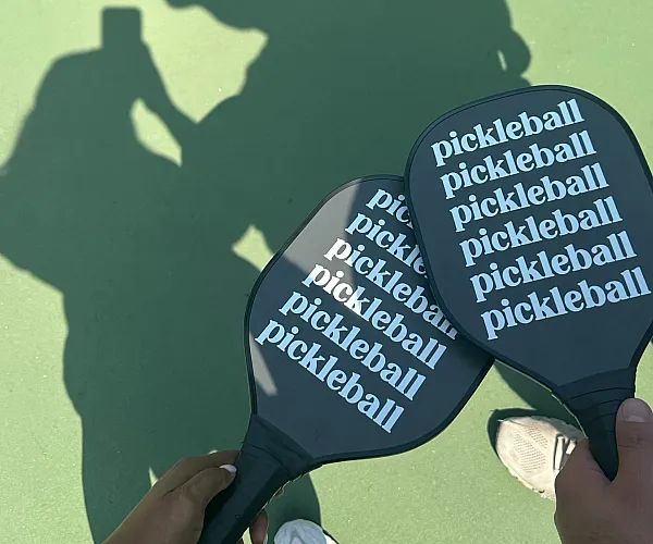 Sunlit pickleball court with players holding paddles casting playful shadows on the green surface.