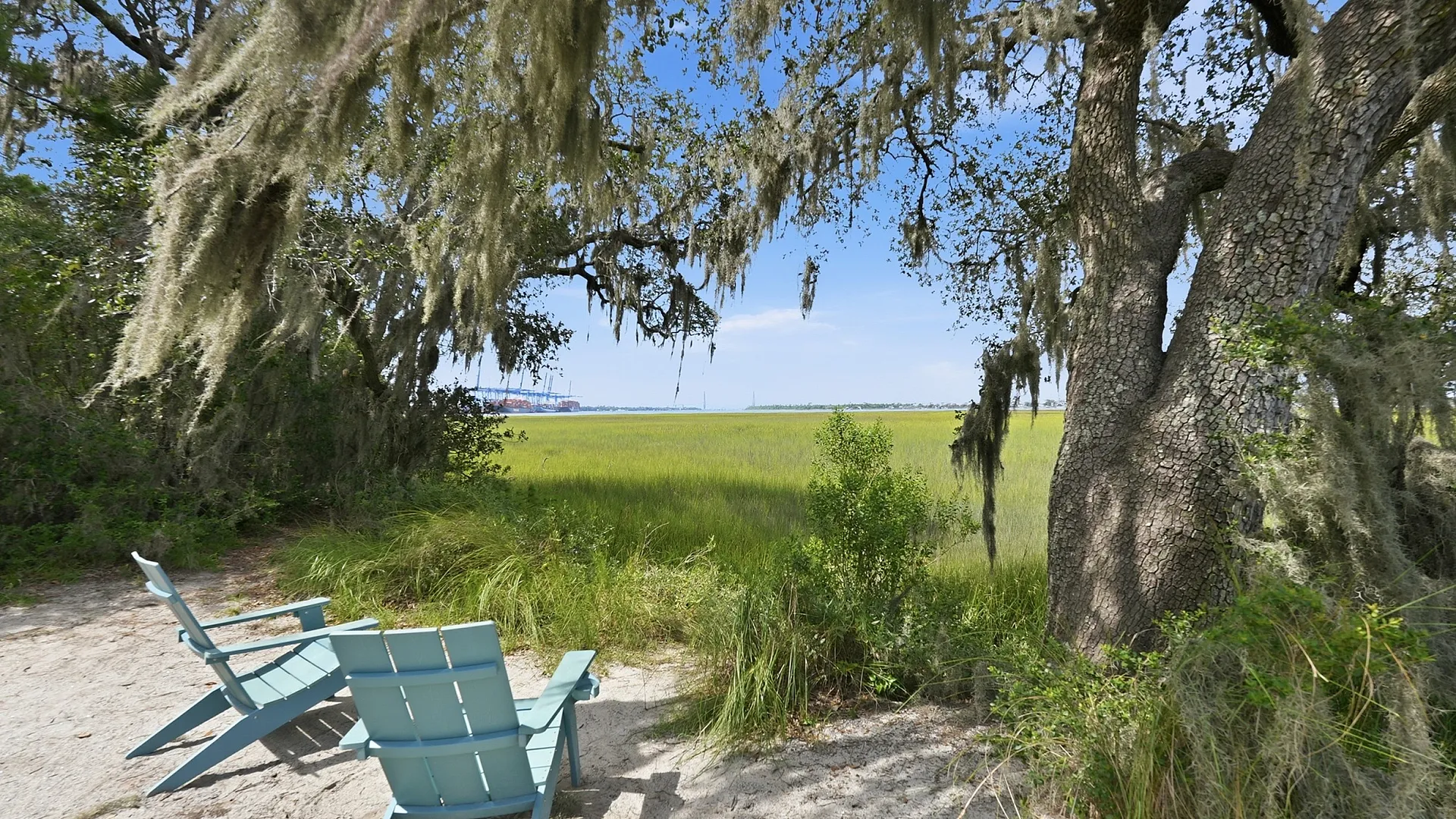 Serene outdoor view featuring two turquoise Adirondack chairs under a picturesque tree draped with Spanish moss, overlooking lush marshland and a distant waterway under a clear blue sky.