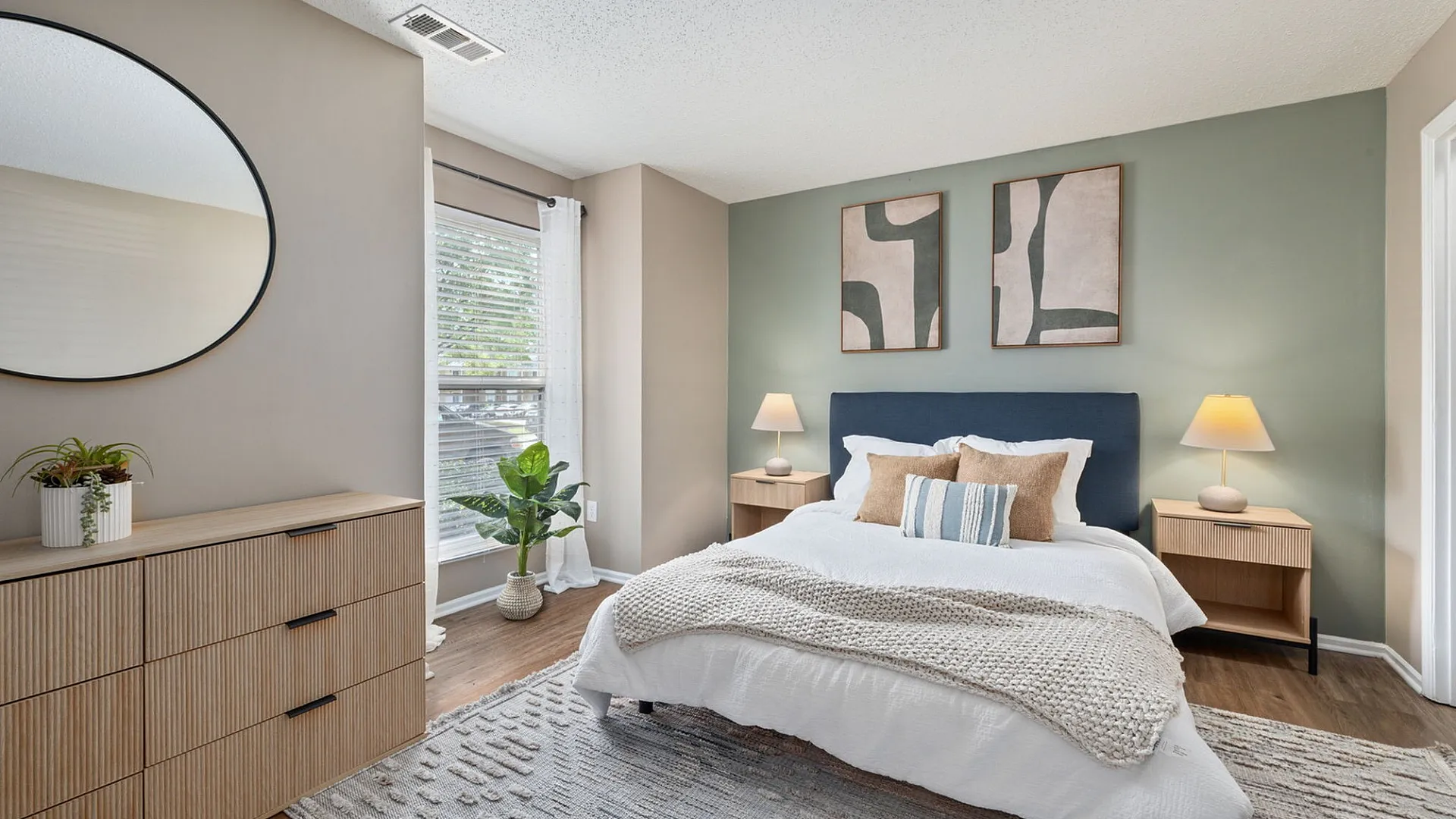 Inviting bedroom with modern decor, featuring a stylish bed with cozy pillows, a large round mirror, and elegant wooden furniture, enhanced by soft natural light.
