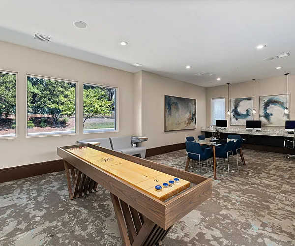 Bright and spacious recreation room featuring a shuffleboard table, comfortable seating, modern artwork, and ample natural light from large windows.