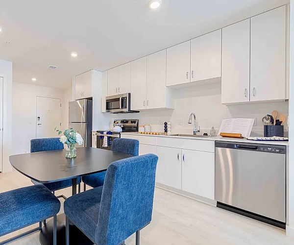 Elegant kitchen and dining area with modern white cabinets, stainless steel appliances, and plush blue chairs around a stylish black table.