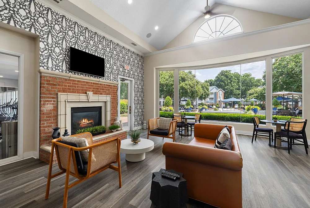 Bright and inviting community lounge featuring modern seating, a cozy fireplace, and expansive windows with a scenic view of lush greenery and outdoor seating.