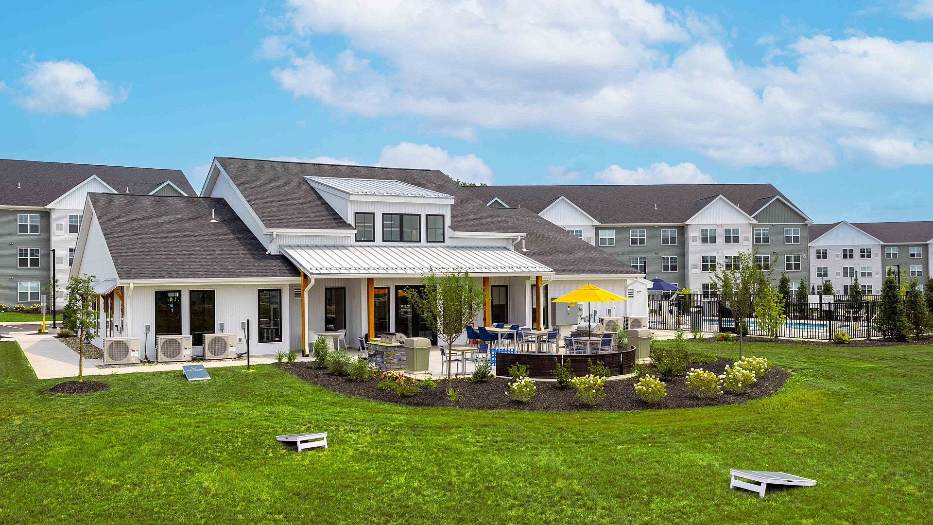 Charming apartment complex with elegant clubhouse, modern outdoor seating area, manicured green lawn, and vibrant community ambiance under a bright blue sky.