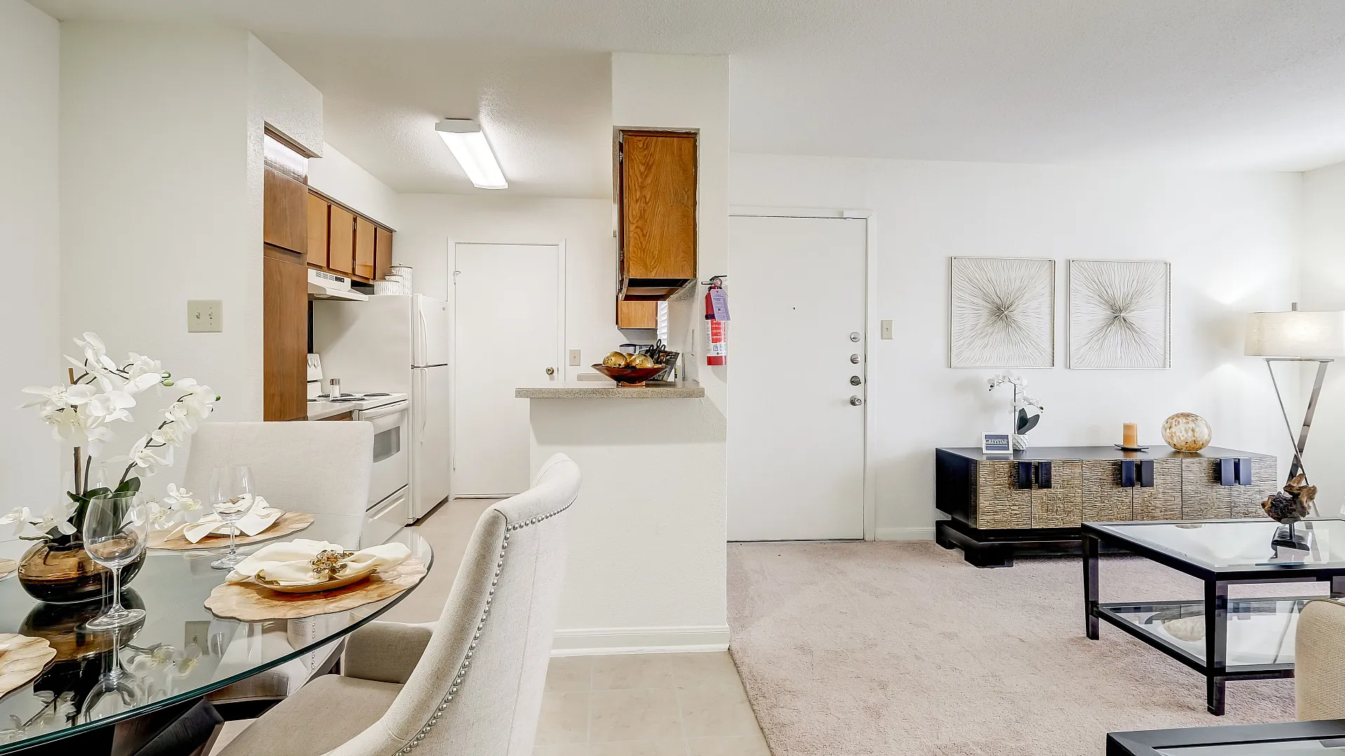 Chic dining area with elegant seating and a glass-top table, adjacent to a cozy kitchen with warm wooden cabinetry.
