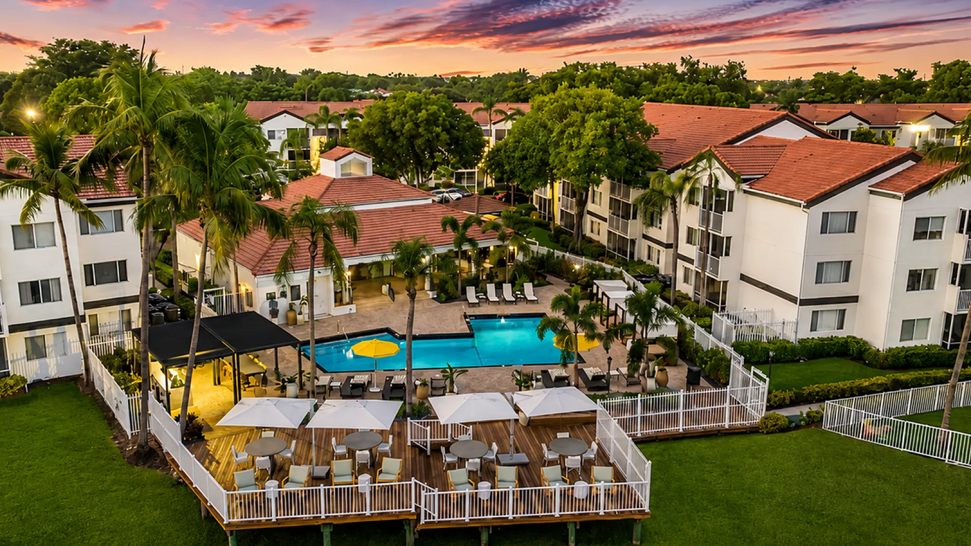 Luxurious apartment complex featuring a stunning pool area, elegant outdoor seating, and elegant architecture amid tropical palm trees at sunset.