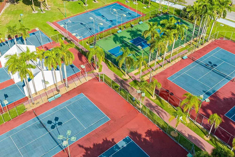 Scenic aerial view of vibrant sports complex featuring multiple tennis and basketball courts surrounded by lush greenery and palm trees.