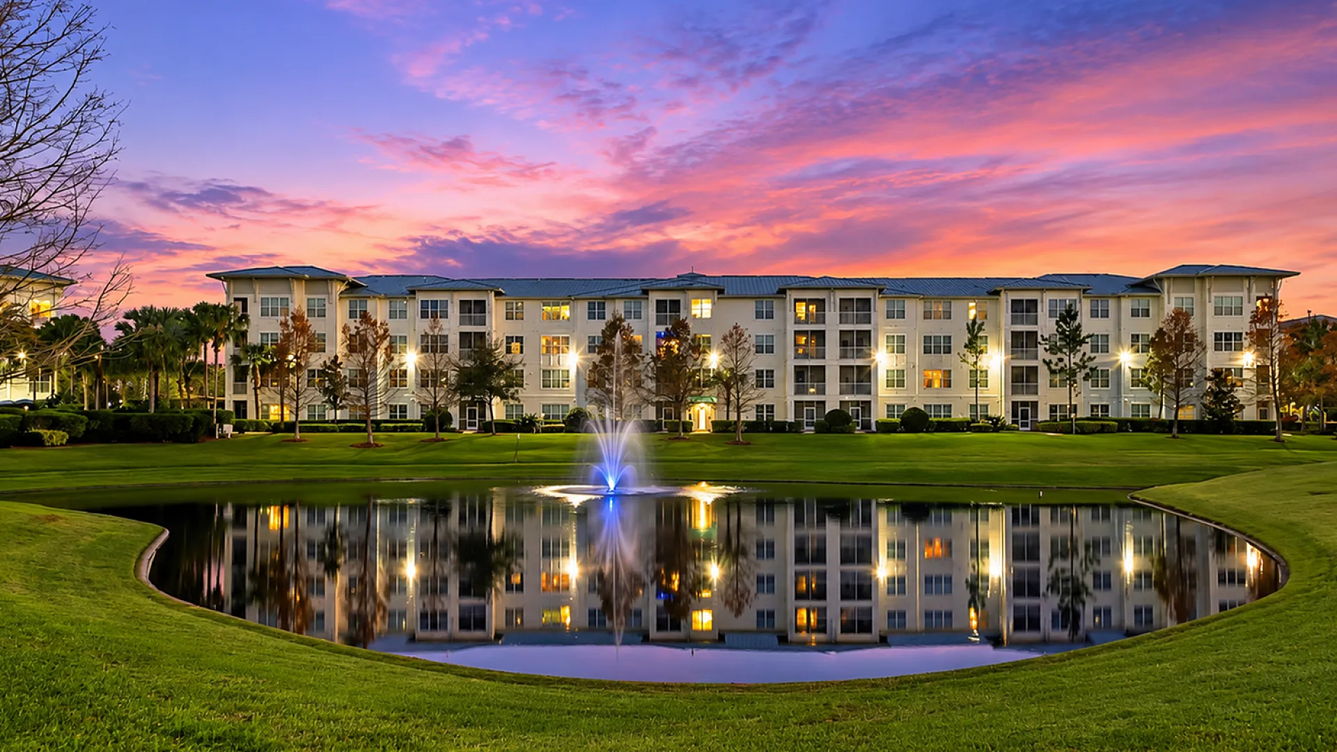 Elegant apartment complex with a serene pond and fountain, highlighted by a stunning sunset sky.