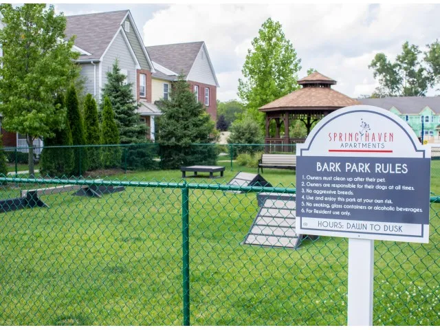 Bark Park fenced in with green grass and some obstacles the animals can use. Green trees surrounding the area with a gazebo in front of the building.