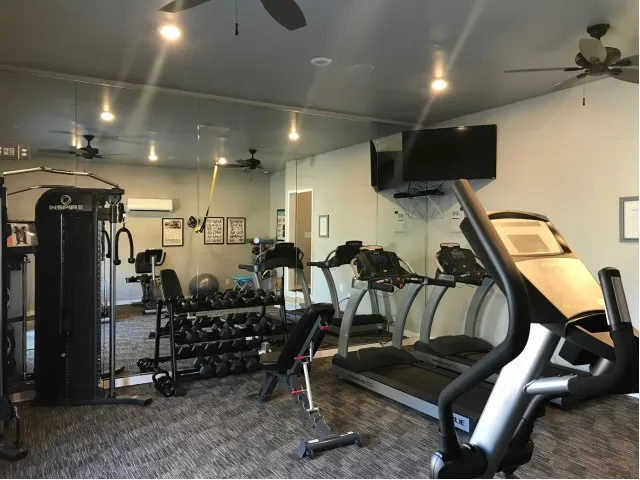 Gym with one wall all mirror, two treadmills, a weight rack, TV in the corner with two ceiling fans. Carpeted area